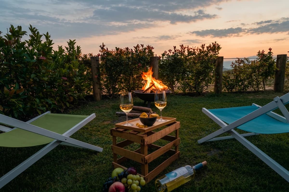 Lounge chairs around a lit fire pit at sunset, with wine and fruit, at The Nest