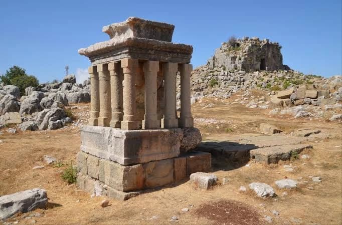 A small columned shrine on its stone base among the limestone boulders at Faqra, a ruined tower behind